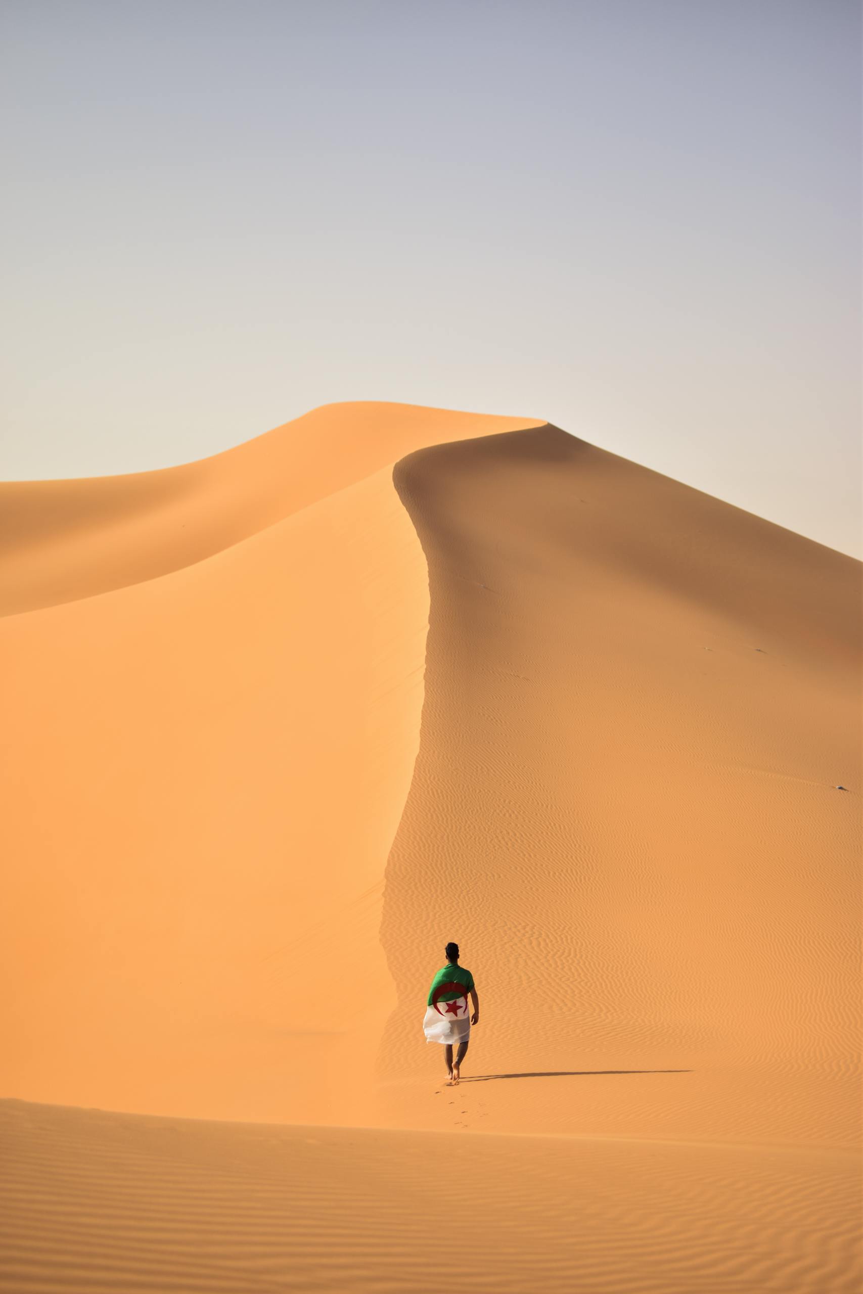 A lone traveler walking over the vast, golden sand dunes of the Sahara Desert in Algeria.