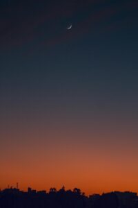 A serene crescent moon in a colorful dusk sky above a city silhouette, capturing twilight's beauty.