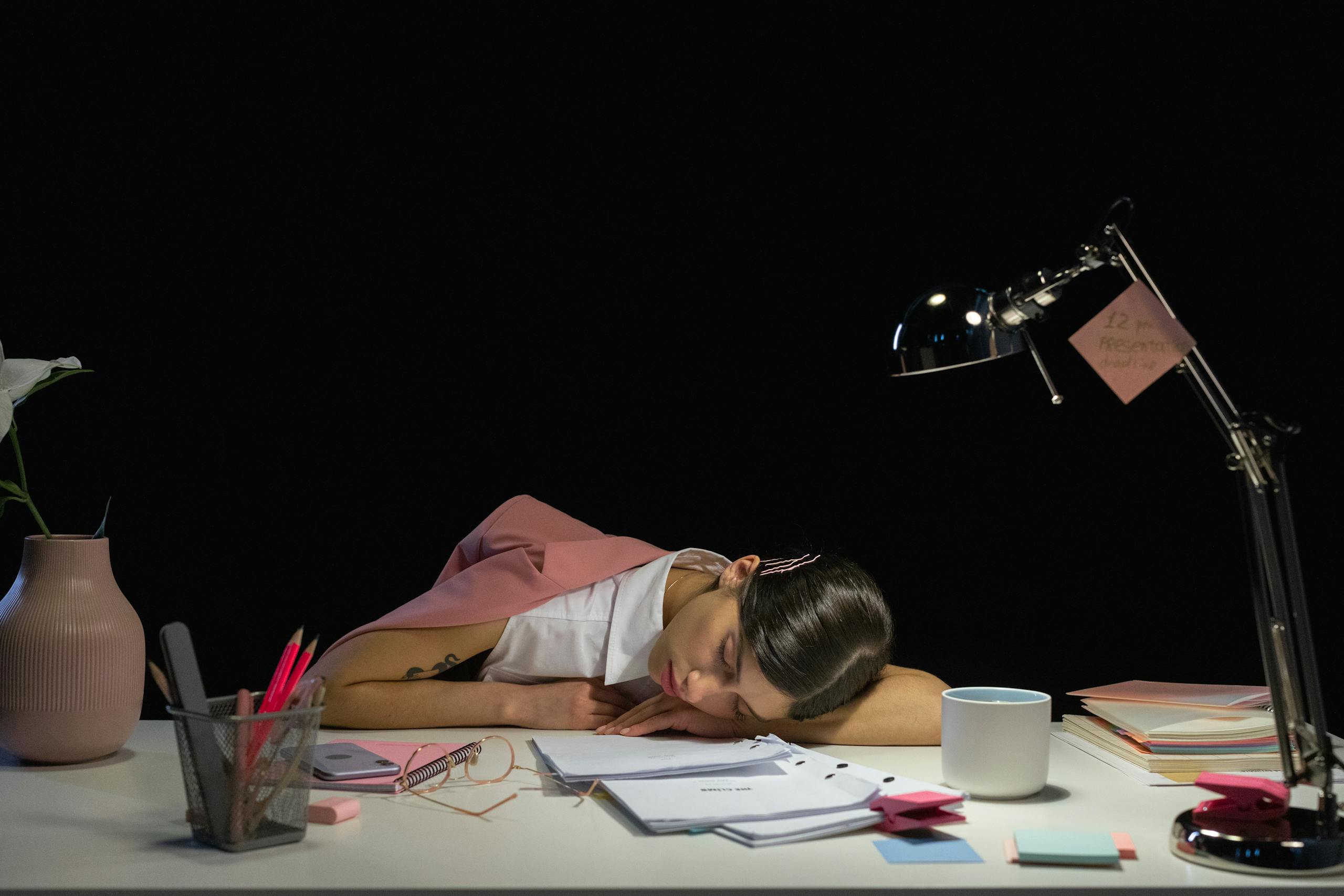 Tired woman asleep on a desk with office supplies under a lamp in dark workspace.